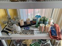 Wide view of assorted candles, multiple candle holders, inspirational plaques, nutcracker tools, and a folded colorful table runner on a white shelving unit.