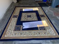 Full view of rectangular navy blue area rug with geometric and floral patterns in beige and gold. Light reflects through garage windows creating bright spots on the rug.