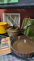 Photo showing yellow cast iron pot, brown cast iron pot with lid, green heating tray, digital thermometer in packaging, and Brown ceramic pie dish.
