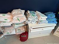 Various folded double size sheets and bedding items stacked on and around a dresser.