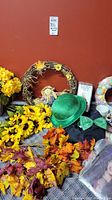 Wide view of fall-themed faux florals including sunflowers, leaves, a fall wreath with a scarecrow figure, and holiday accessories such as green hat and bow tie.