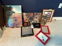 Collection of assorted picture frames and wall plaques arranged on floor against wall. Includes large footprint image plaque, various wood and plastic frames, and Tic Tac Toe decor.