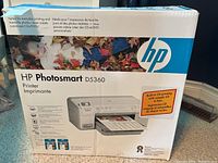 Front of box displaying HP Photosmart D5360 printer image, product details with a photo of a child, and listing features including built-in CD printing.