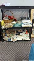 Two shelves filled with assorted folded linens including placemats, napkins, tablecloths, basket with some small cloth items, mixed patterns and plain.