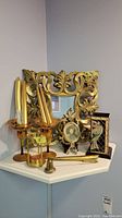 Wide-angle photo showing gold themed decor items arranged on a white corner shelf: ornate gold mirror, three tall gold candle holders with taper candles, small decorative gold picture frame, small gold bell, and small gold hourglass with orange sand.