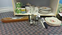 Wide view showing mugs, plates, saucers stacked on a shelf, wooden handled knives in a holder on the floor, metal salt and pepper shakers with stand, and various utensils.