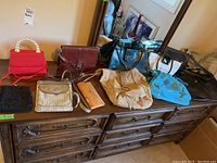 Photo showing nine assorted purses arranged on a wooden dresser including fabric and leather bags in different colors and sizes.