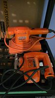 Orange Black and Decker sander and jig saw placed on a black plastic shelf. Power cords are wrapped around each tool.