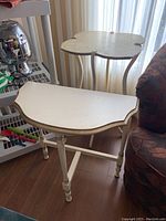 Two painted wood side tables shown together. The smaller two-level table has a clover-shaped top with curved legs. The larger single-level table has a scalloped edge top with turned legs. Both painted white with wear and some water stains visible.
