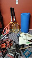 Photo of all items together: yoga mat rolled up, two Ironman racquets, white ice skates size 6, weights, rubber bands on carpeted floor against red wall