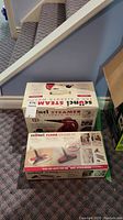 Two boxed items side by side: one handheld steam cleaner, one floor steamer kit attachment
