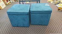 Front view of two blue square storage ottomans placed side by side on carpeted floor.