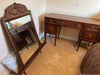 Wood dressing table and matching freestanding mirror placed against a beige wall with carpeted floor. Mirror shows carved detail at top. Table has five drawers with brass ring pulls and slightly curved legs.