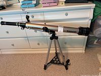 Front and side view of the Tasco Luminova telescope on its tripod stand against a white dresser background.