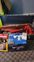 Overall view of black metal toolbox containing a red caulking gun, wooden level, boxed Heath-Zenith motion sensor light, and boxed Rayovac headlamp.