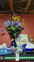 Photo showing cobalt blue glass vase filled with multi-colored faux flowers including purple, yellow, and green artificial greenery, along with plates, a bowl, clown figurines, and placemats arranged on a shelf.