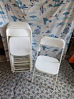 Photo showing a stack of 9 white folding chairs and one unfolded chair in front, against a patterned backdrop.