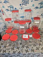 18 clear plastic containers arranged in three rows with red lids. 16 smaller square containers in the front and middle row, 2 larger square containers in the back row. Containers used for sorting Lego pieces.