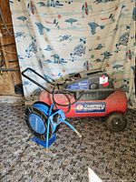 Full view of red Campbell Hausfeld air compressor with blue hose reel in front, placed on floor with patterned fabric backdrop showing Star Wars design.