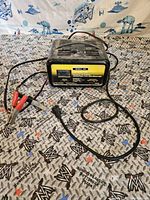 Front view showing battery booster and charger with cables and clamps on a patterned surface with Star Wars themed background.