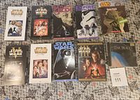 Top view of all 10 items laid out: 7 VHS cases, 1 DVD case, 1 book, 1 CD case