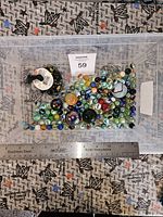 Top view of clear plastic container filled with multiple assorted vintage marbles of different sizes and colors on patterned fabric background. Stainless steel ruler placed in front for scale.