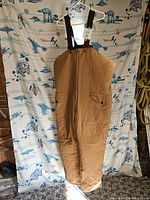 Brown winter overalls hanging on a hanger, showing full front view with flap pockets and black adjustable suspenders