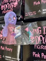 Top view showing a single Living Dead Dolls Pink Posey figurine with purple hair, pink dress, pale face with wide eyes, and a pink rose accessory in a plastic bag, on top of multiple original black boxes.