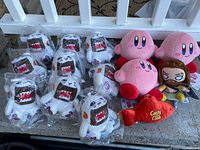 Photo showing 14 plush toys including multiple Kirby and Cat Domo plush toys, one warrior plush toy, and a Candy Crush plush toy in original plastic packaging.