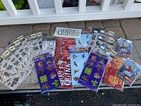 24 packages of new sealed sticker and temporary tattoo sets arranged outdoors on floor; includes Batman, League of Legends, Suicide Squad, Marvel Avengers, The Walking Dead themes.