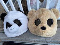 Two plush adult-sized head masks: one beige bear and one white panda with black facial markings, seated outdoors on a step.