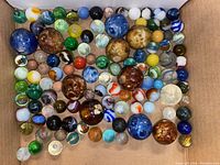 Overhead image of a large variety of vintage glass marbles arranged in a cardboard box, showing multiple colors, sizes, and patterns.