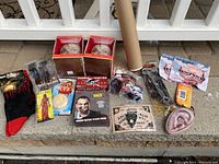 Lot of Halloween and horror themed items displayed on pavement including two horror skull masks in boxes, a rolled Nightmare on Elm Street poster, various small collectibles including zombie hunter, Ouija board, coasters, Deadpool keychain, novelty ear prop, and snack bar.