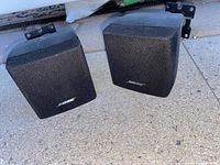 Pair of Bose cube mini speakers with attached wall mounting brackets placed on an outdoor concrete surface. View shows front and side of speakers.