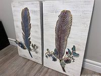 Two metal accent wall art panels displayed side by side. Each panel features a large feather with three-dimensional floral and leafy metal accents on a painted white textured background.