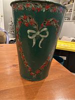 Green hand-tole painted sap bucket with heart wreath and white bow on wooden table, showing decorative painted details.