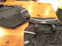 Two black Axiom saddlebags placed on wooden surface showing front and side views with logo, reflective striping, mesh pocket, and buckles visible.