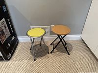 Photo of two collapsible stools side by side on carpet. One stool wooden seat, black legs; other stool yellow seat, metal frame.