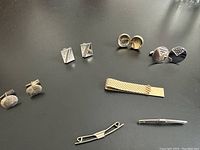 Four pairs of French cufflinks, three tie clips, and one money clip arranged on a dark surface.