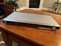 Photo of silver Daytek DVD player placed on a wooden table, showing front view with disc tray and power button.