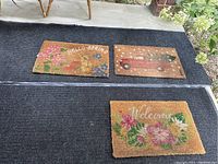 Three outdoor coir mats including 'HELLO SPRING' with flowers, red vintage truck, and 'Welcome' with pink flowers, on top of a dark gray runner rug.