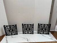 Three square metal wall art panels with black ornate floral and swirl pattern over silver backing arranged side by side on floor.