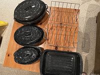 Four black speckled roasting pans, three with lids and one without, and two wire turkey racks on wooden table.