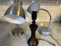 Three lamps on countertop: stainless steel lamp, brown wooden lamp base without shade, and periwinkle desk lamp with vented head.