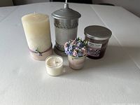 Five assorted used candles are displayed on a light surface. The candles include a large ivory pillar candle with a purple string tie, a silver metal container candle with a handle, a small purple jar candle labeled 'Trendy Home', a white tealight candle, and a decorative pastel flower candle.