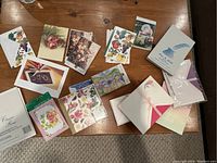 Table photo of assorted blank notecards and writing paper packages, with floral, fruit, and decorative designs, mostly unopened packages.