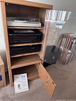 Full frontal view of the audio stereo cabinet showing glass door upper section and wood door lower section, both doors open exposing interior shelves and instruction manual placed on floor.