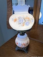 Full view of the hurricane lamp showing the floral painted glass globe and base along with the ruffled glass chimney top and metal ornate base.