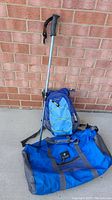Full lot showing the blue hydration backpack, blue Outdoor Products duffle bag, and Swissgear walking stick leaning against a brick wall