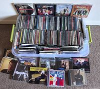 Plastic storage bin filled with a large collection of music CDs organized upright, with some CDs displayed in front on carpeted floor. Visible artists include Natalie Merchant, Adam Sandler, Glenn Miller, Buddy Holly, and others.